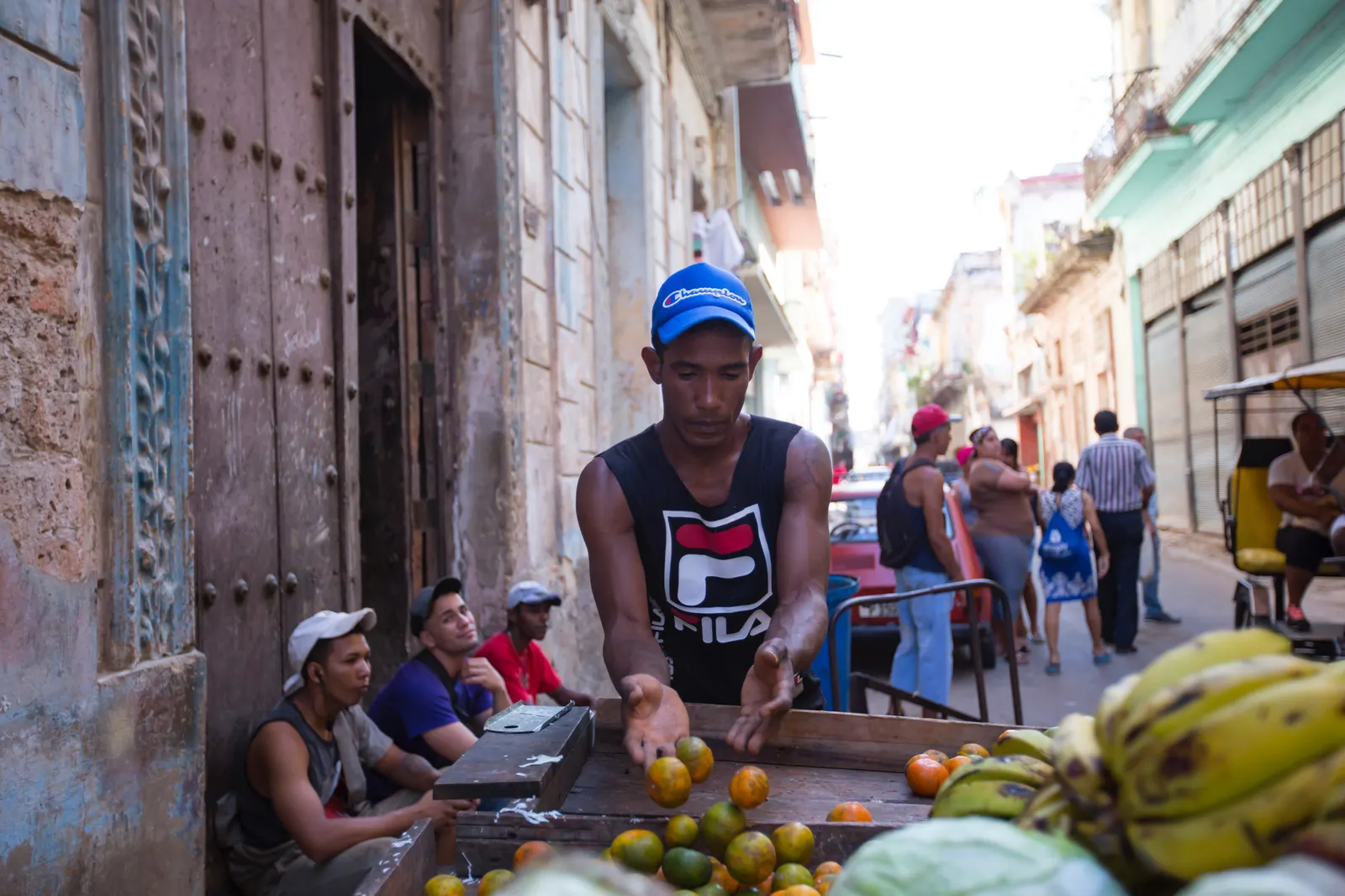 A young man in a baseball cap sorts oranges on a wooden cart in a narrow street, neighbors sitting in a doorway behind him.