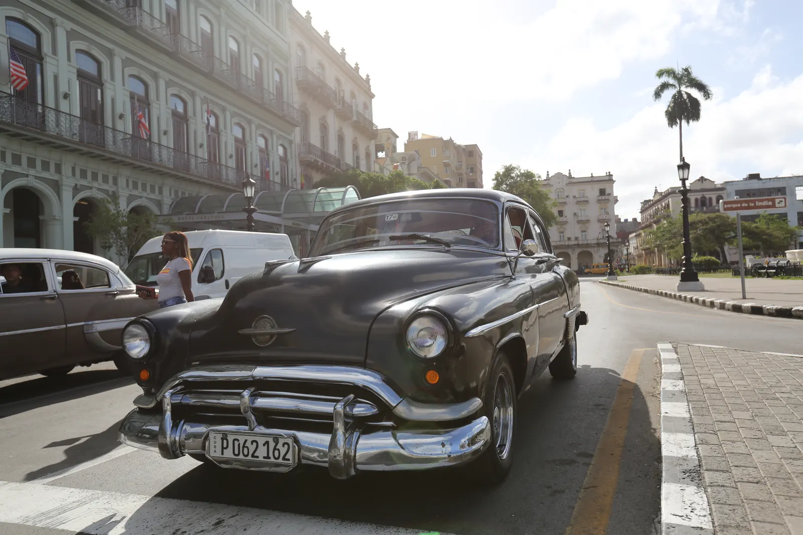 A black 1950s Oldsmobile parked on a sunlit boulevard in front of a long colonnaded hotel.