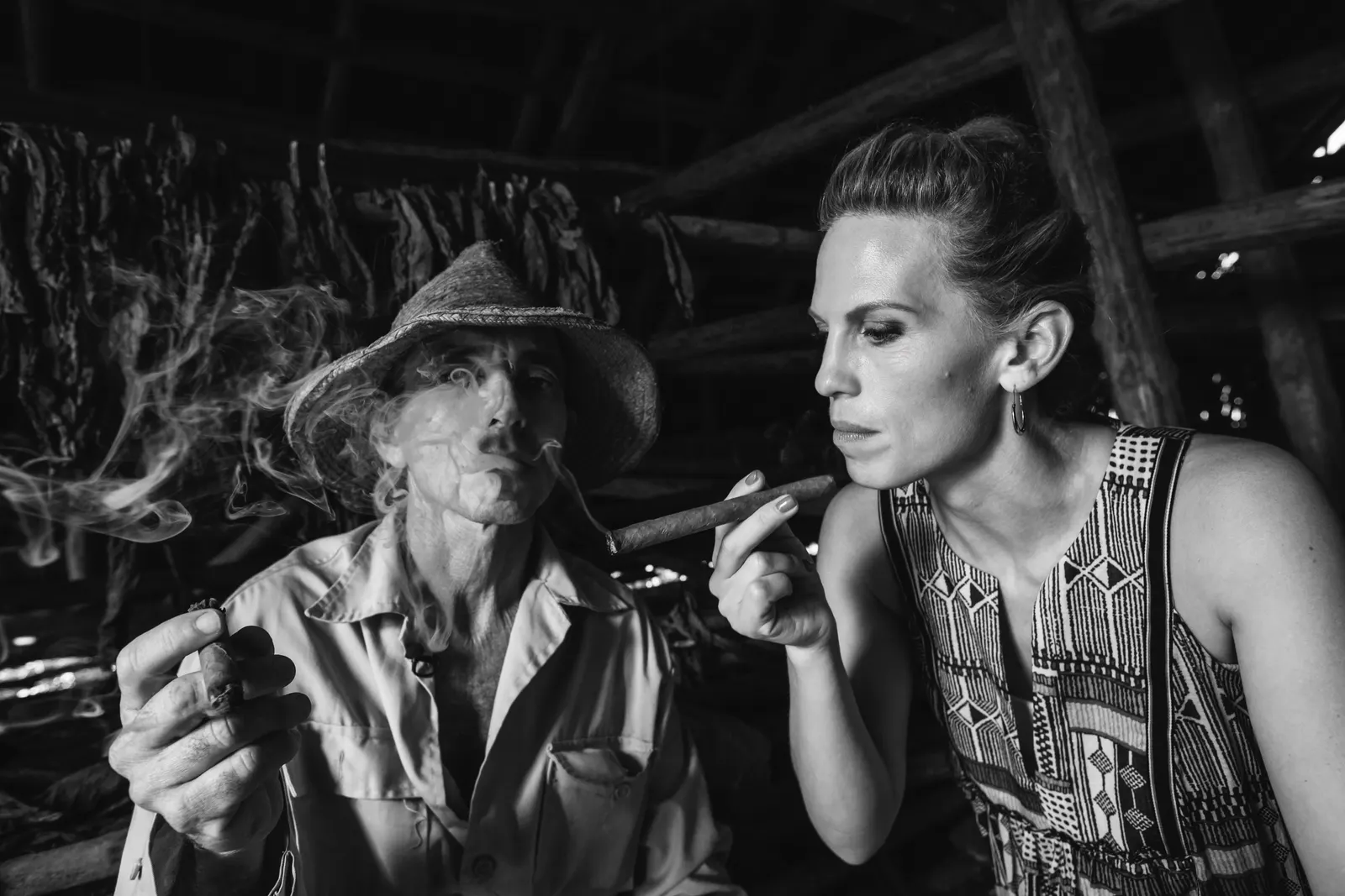 Black and white: a tobacco farmer in a straw hat lights a cigar for a woman inside a drying barn, rows of curing leaves hanging behind them.