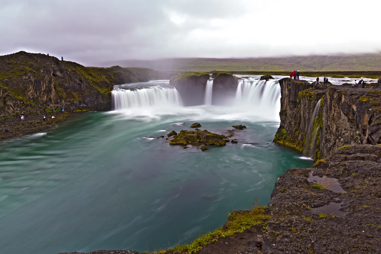 A wide horseshoe-shaped waterfall under a low gray sky, turquoise water running past mossy black rock.