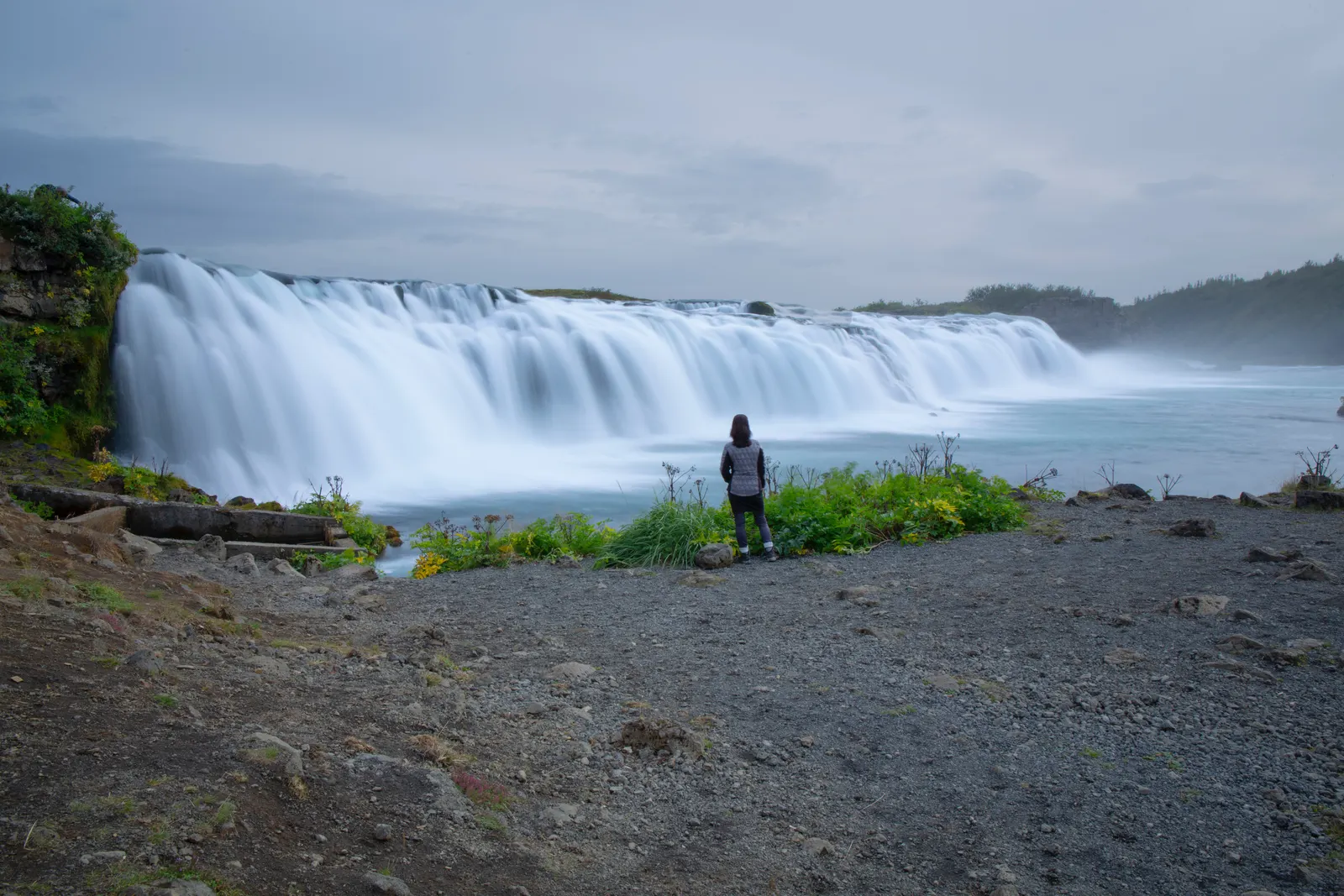 A person stands alone on a gravel bank facing a broad curtain of falling water.