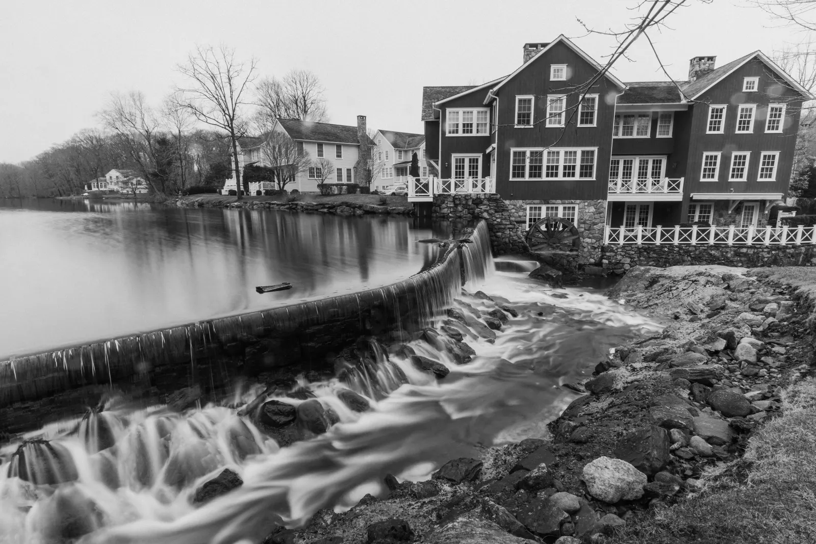 Black and white: water spilling over a curved dam below a clapboard mill building at the edge of a still pond.