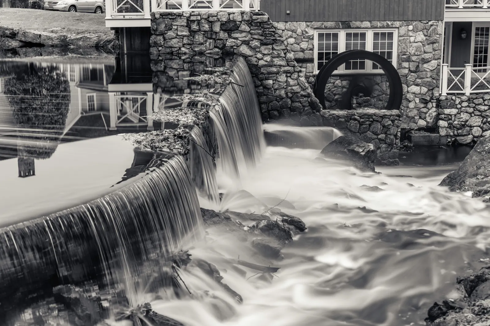 Black and white: a long exposure of water pouring over a stone spillway beside a mill's iron waterwheel.